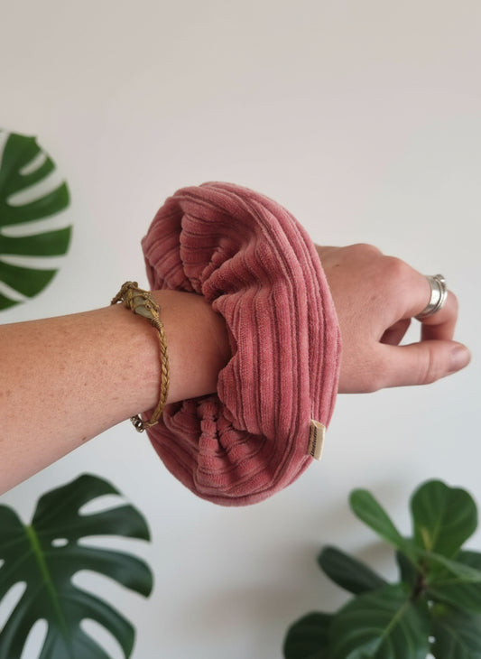 Hot pink handmade cordury chunky scruchie, complete with custom organic cotton Aurora's Destination tag sewn into the seam. This scrunchie looks soft and thick. It is placed on a womans arm, next to a crystal macrame bracelet. The woman is wearing silver rings and in the background we can see a few plants.