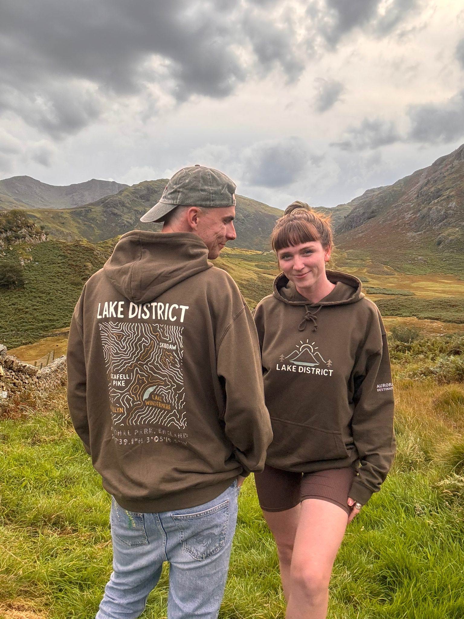 Two people wearing matching olive-green Lake District hoodies stand in a scenic mountain landscape. The woman faces the camera with a smile, while the man shows the back of his hoodie, which features a topographic map design. Olive / earth green organic hoodie. Sustainbly made with triple prints. A sleeve print, front print and large back print. Featuring earthy tones of the Lake district map, co-ordinates and the popular lakes - Lake Windermere, Scarfel Pike, Skiddaw and Helvellyn. Cute outdoorsie couple.