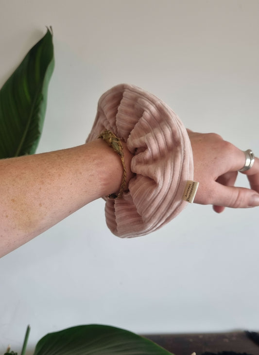 Rose pink handmade cordury chunky scrunchie, complete with a custom organic Aurora's Destination tag sewn into the seam. Scrunchie is on a womans writst, next to a crystal macrame bracelet. She's also got silver rings on. There are plants in the background.