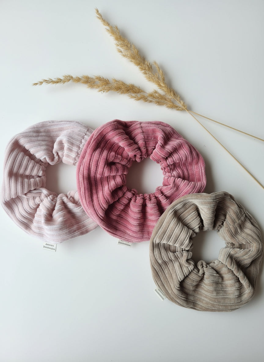 3 handmade cordury chunky scrunchies. Hot pink, rose pink and natural beige. All complete with a custom organic cotton Aurora's Destination tag. Accessories on a smooth white surface, with two dried flowers to the side. 
