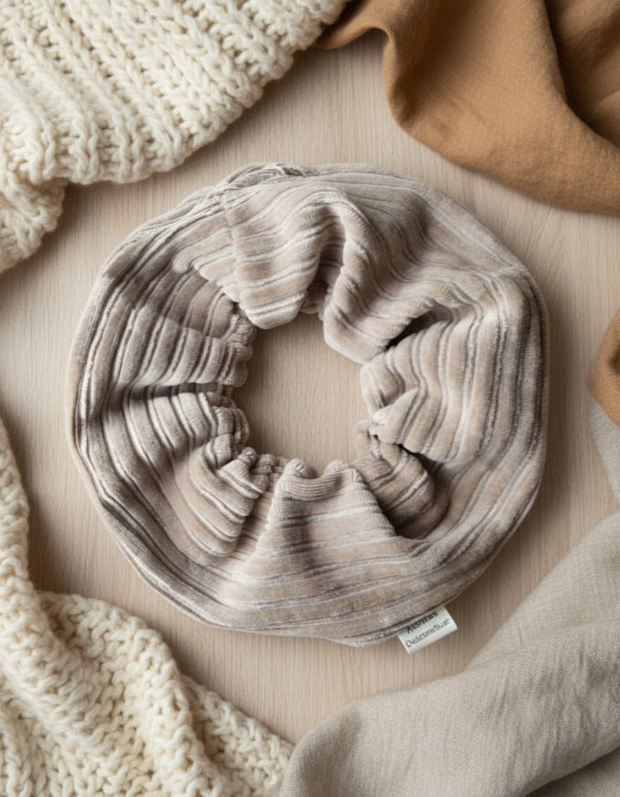 Natural Beige cordury handmade scrunchie, thick and chunky in shape. With a custom cotton Aurora's Destination tag sewn into the seam. On a cosy fabric background, surrounded by soft nutral tones in a verity of fabrics.