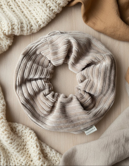 Natural Beige cordury handmade scrunchie, thick and chunky in shape. With a custom cotton Aurora's Destination tag sewn into the seam. On a cosy fabric background, surrounded by soft nutral tones in a verity of fabrics.