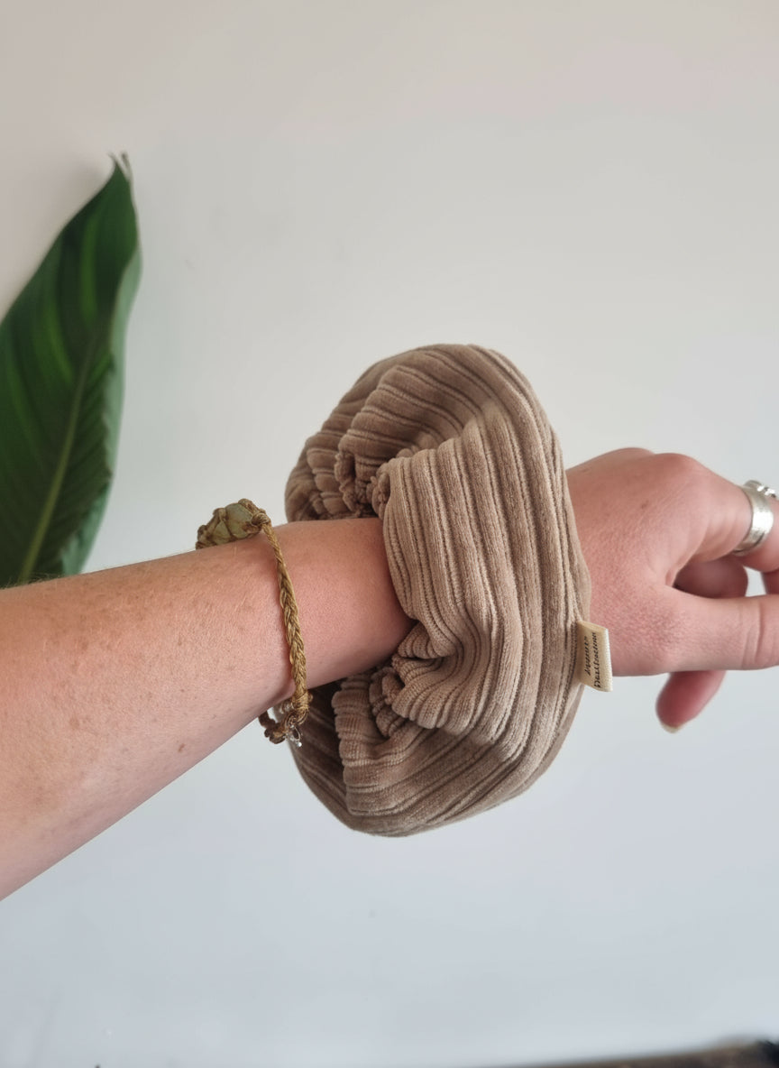 Natural beige cordury chunky scunchie on a womans wrist. Scunchie is handmade with a custom cotton Aurora's Destination tag sewn into the seam. In the image, you can also see the woman is wearing a handmade crystal bracelet, silver rings and there is a banana plant in the background. Minamilis aesthetic, plant lover. Simple life.