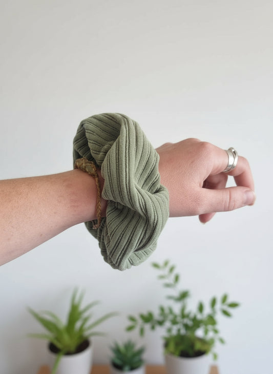 Sage green chunky cordury scrunchie on a womans wrist, with it's custom organic cotton mini tag, reading Aurora's Destination sewn into the seam. The woman is wearing a cystal bracelet and silver rings. Plants can be seen in the background.