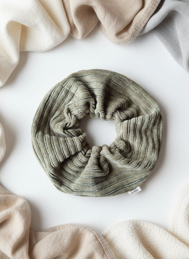 Sage Green Chunky Scrunchie, on a fabric display surrounded by neutral toned organic fabrics. Scrunchie is complete with a custom Aurora's Destination mini organic cotton tag sewn into the seam.