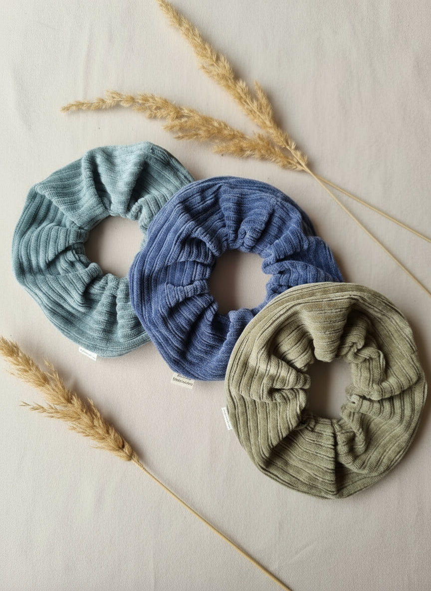 A selection of three handmade chunky scrunchies, complete with organic cotton custom tags reading 'Aurora's Destination' The three scrunchies are placed neatly next to eachother in a curve in the order; blue sage, washed navy then light moss green. They're on a neutral coloured fabirc with a handful of dried flowers and grains scattered around. Aurora's Destination's products are sustainably made, unique, eco friendly and are inspired by nature. These are a great accessory and gift. 