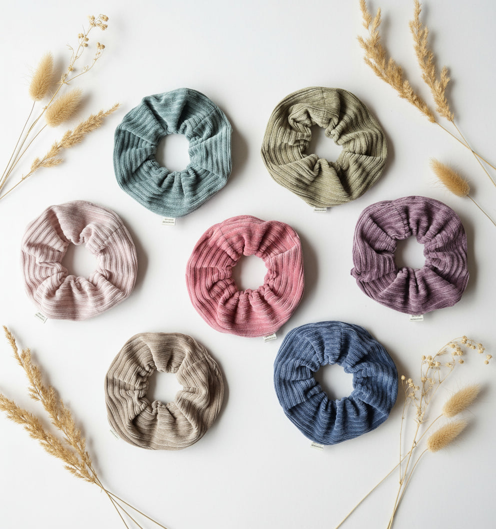 All seven colours of the handmade cordury chunky scrunchie, complete with custom Aurora's Destination organic cotton tag. Aesthetically lay out on a white background surrounded by soft dried flowers.