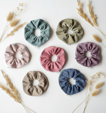 All seven colours of the handmade cordury chunky scrunchie, complete with custom Aurora's Destination organic cotton tag. Aesthetically lay out on a white background surrounded by soft dried flowers.
